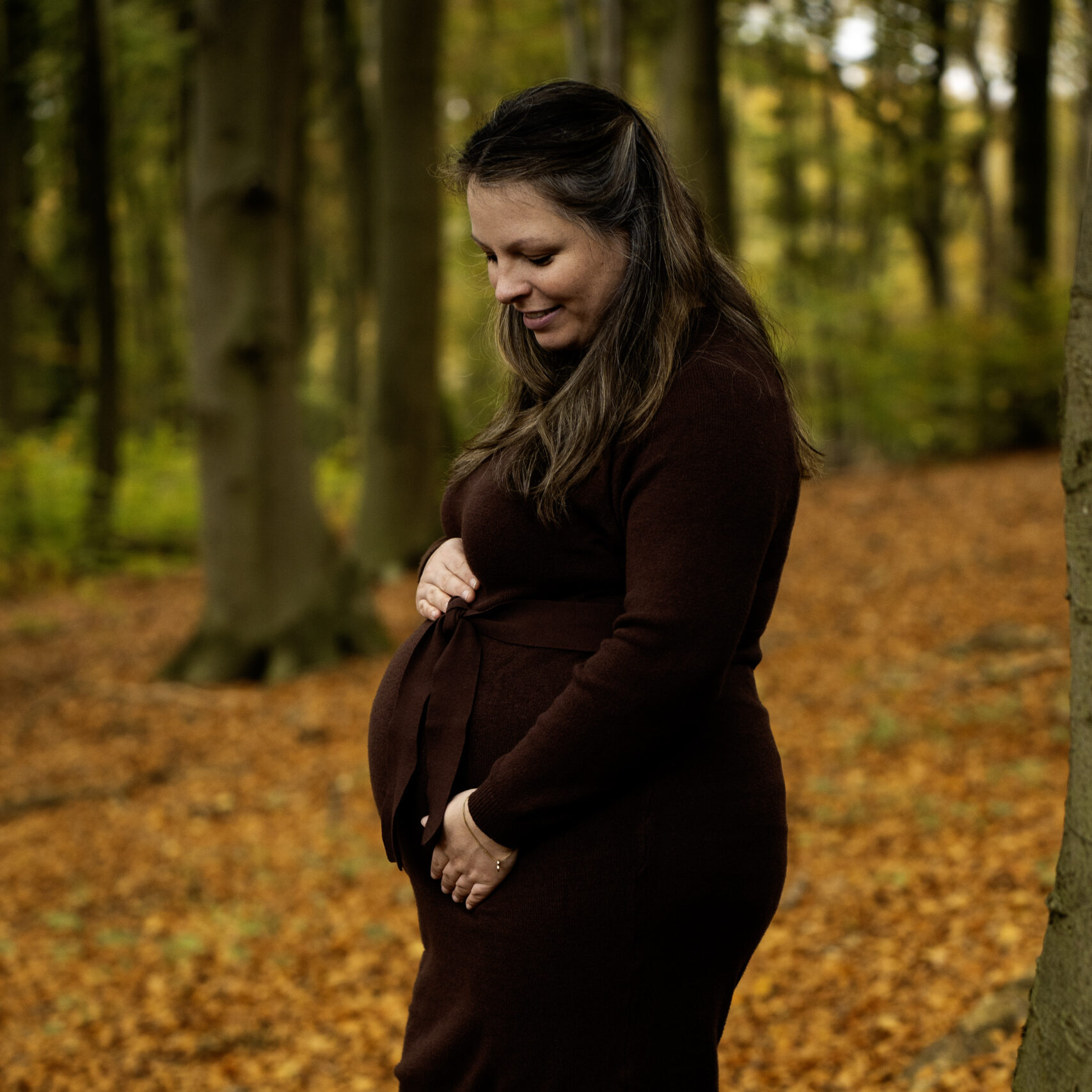 Schwangere Frau im braunen Kleid hält ihren Babybauch und lächelt im Herbstwald.