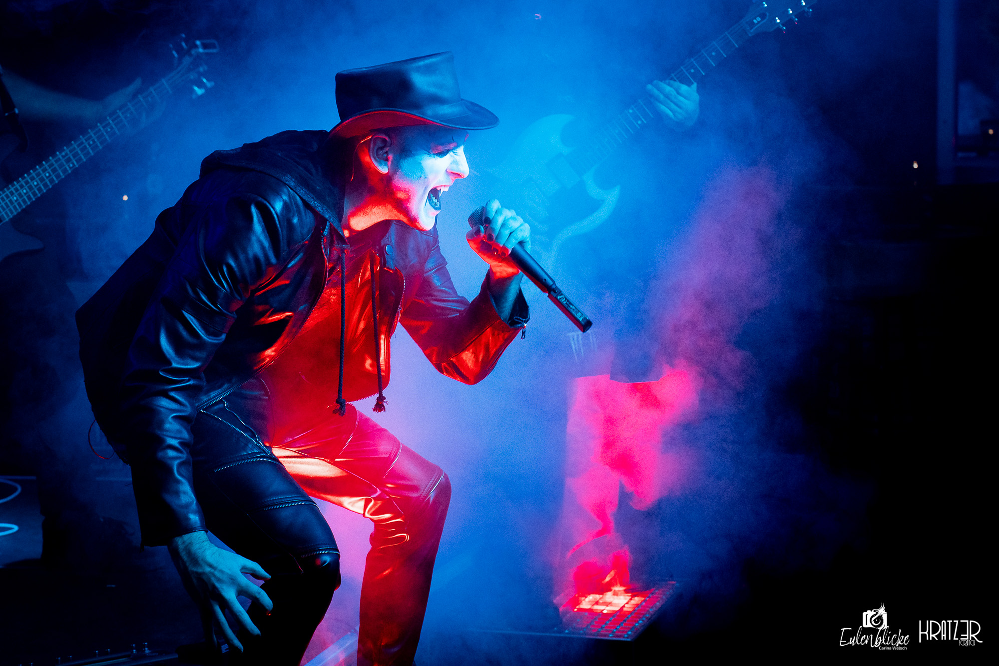 Sänger mit schwarzem Hut geht beim Singen in die Hocke, umgeben von blau und rot beleuchtetem Bühnennebel.