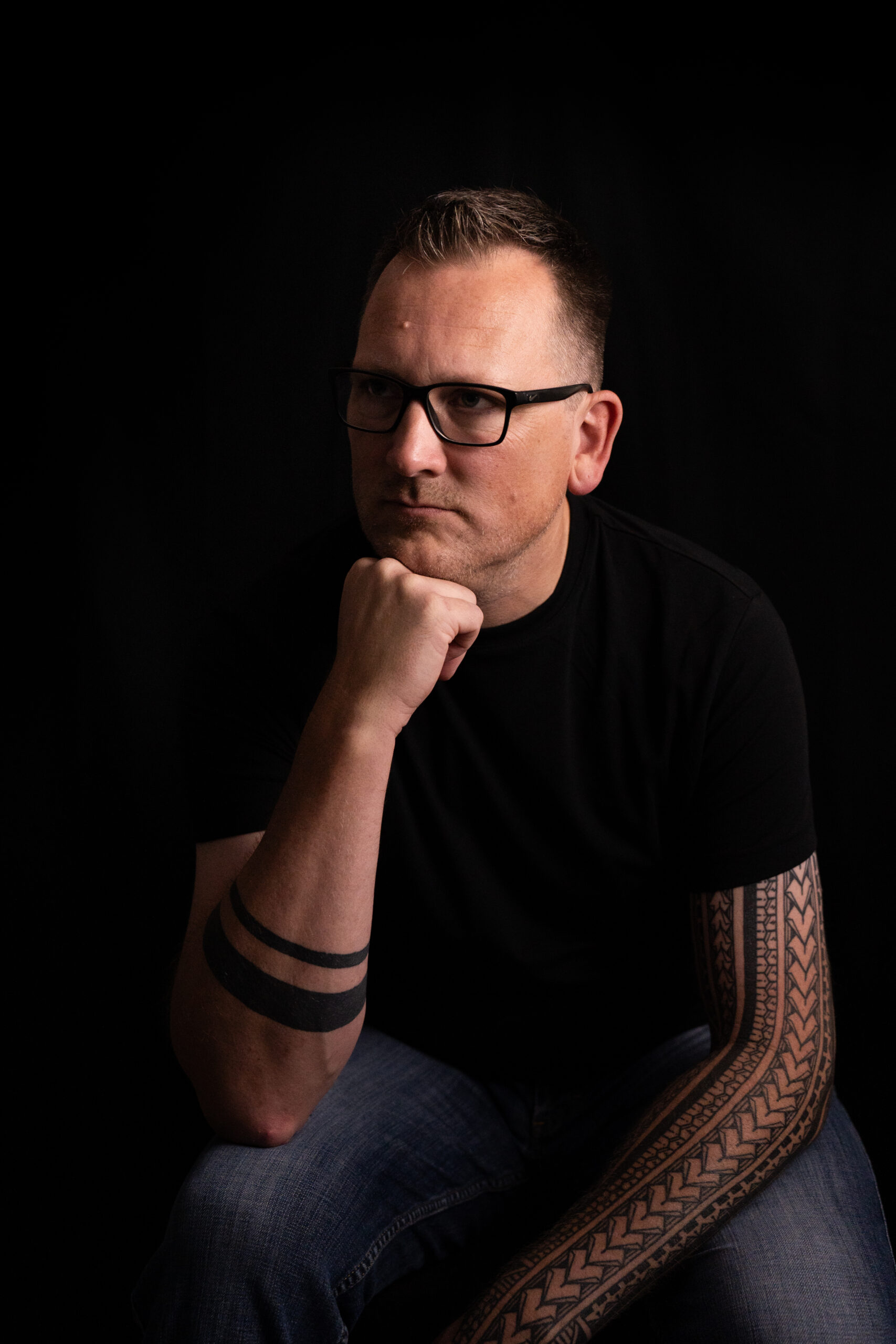 Mann mit Brille stützt sein Kinn auf die Faust und blickt vor dunklem Hintergrund zur Seite.