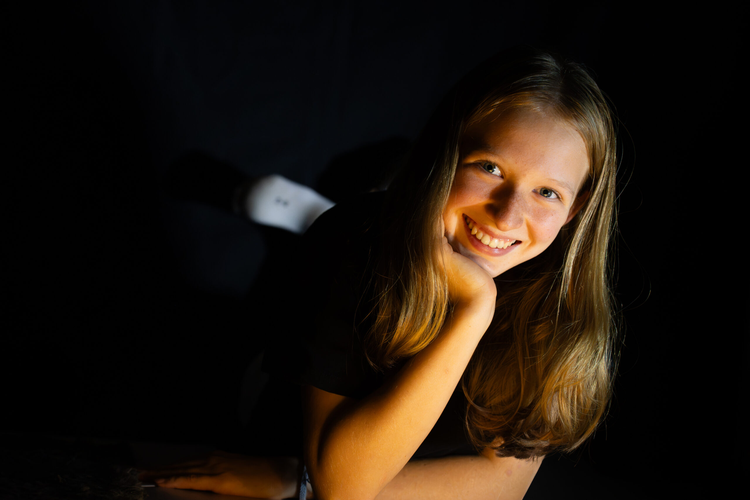 Mädchen legt eine Hand an die Wange und lächelt bei warmem Licht vor dunklem Hintergrund.