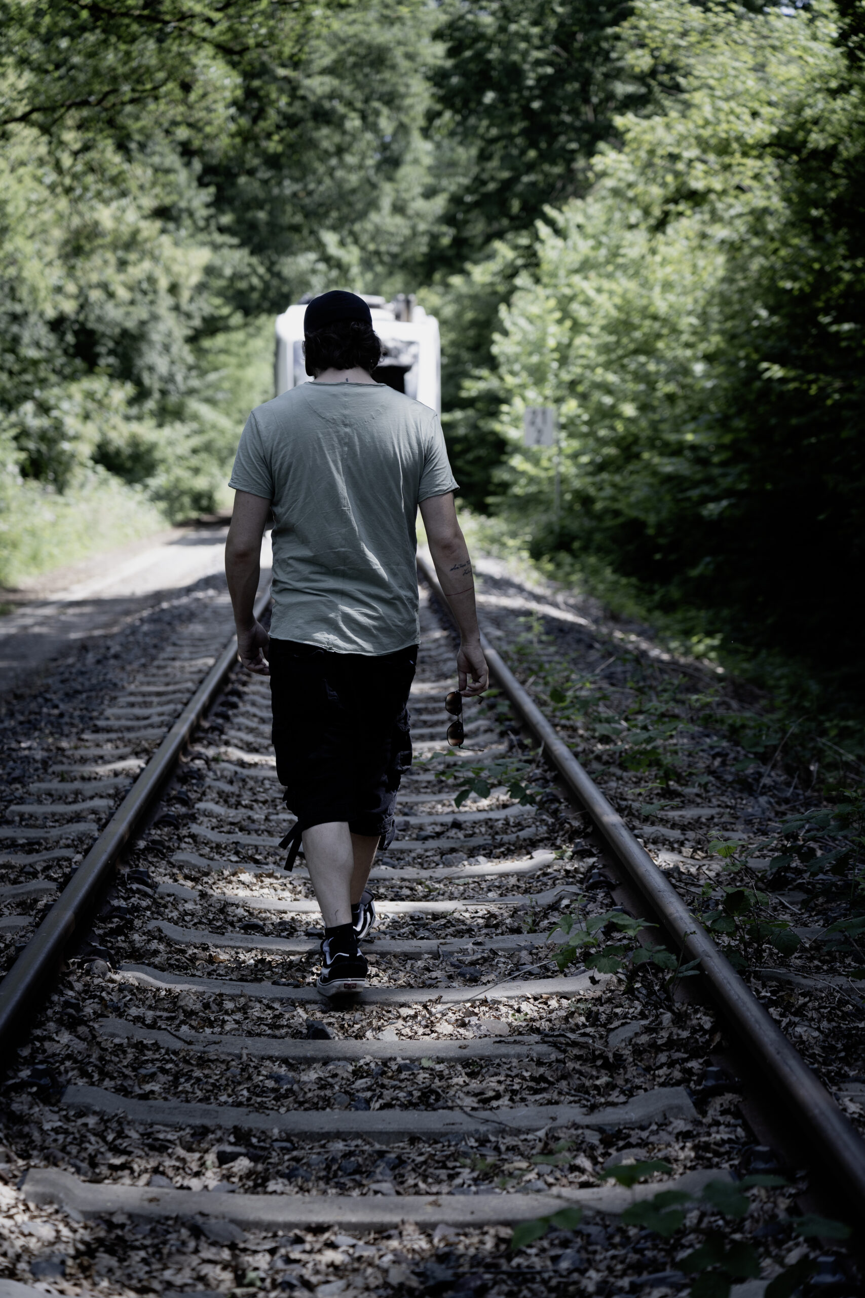 Mann mit Kappe und graugrünem T-Shirt geht auf Bahngleisen zwischen Bäumen von der Kamera weg.
