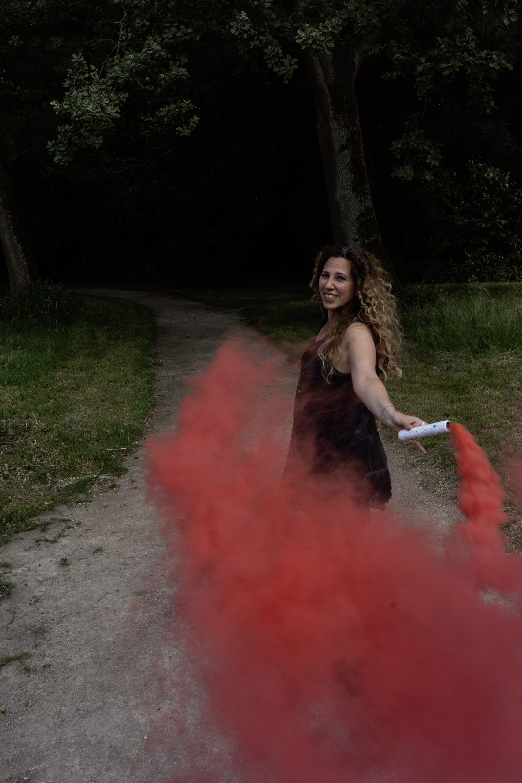 Frau in schwarzer Kleidung blickt auf einem Waldweg zur Kamera, während roter Rauch um sie aufsteigt.